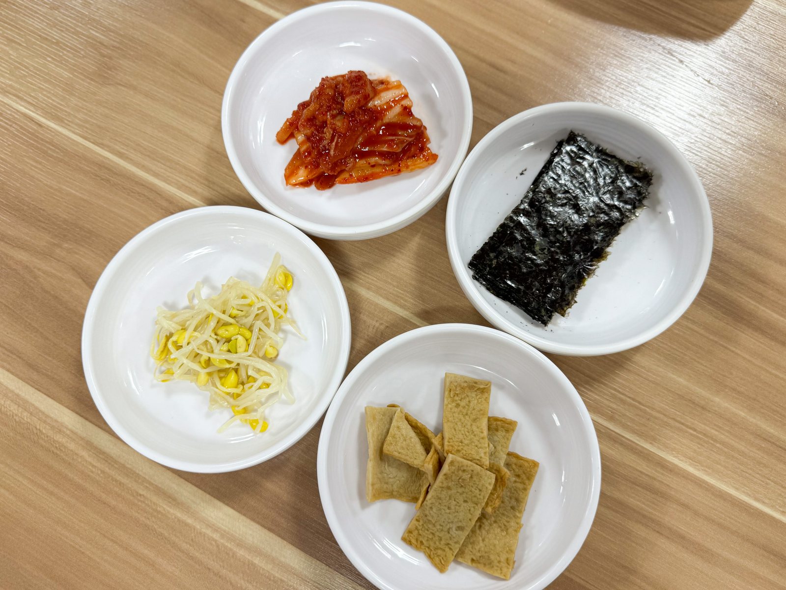 성심조림 제주공항점 기본 반찬 — 밝은 원목 식탁 위 배추김치, 김, 콩나물무침, 어묵볶음.