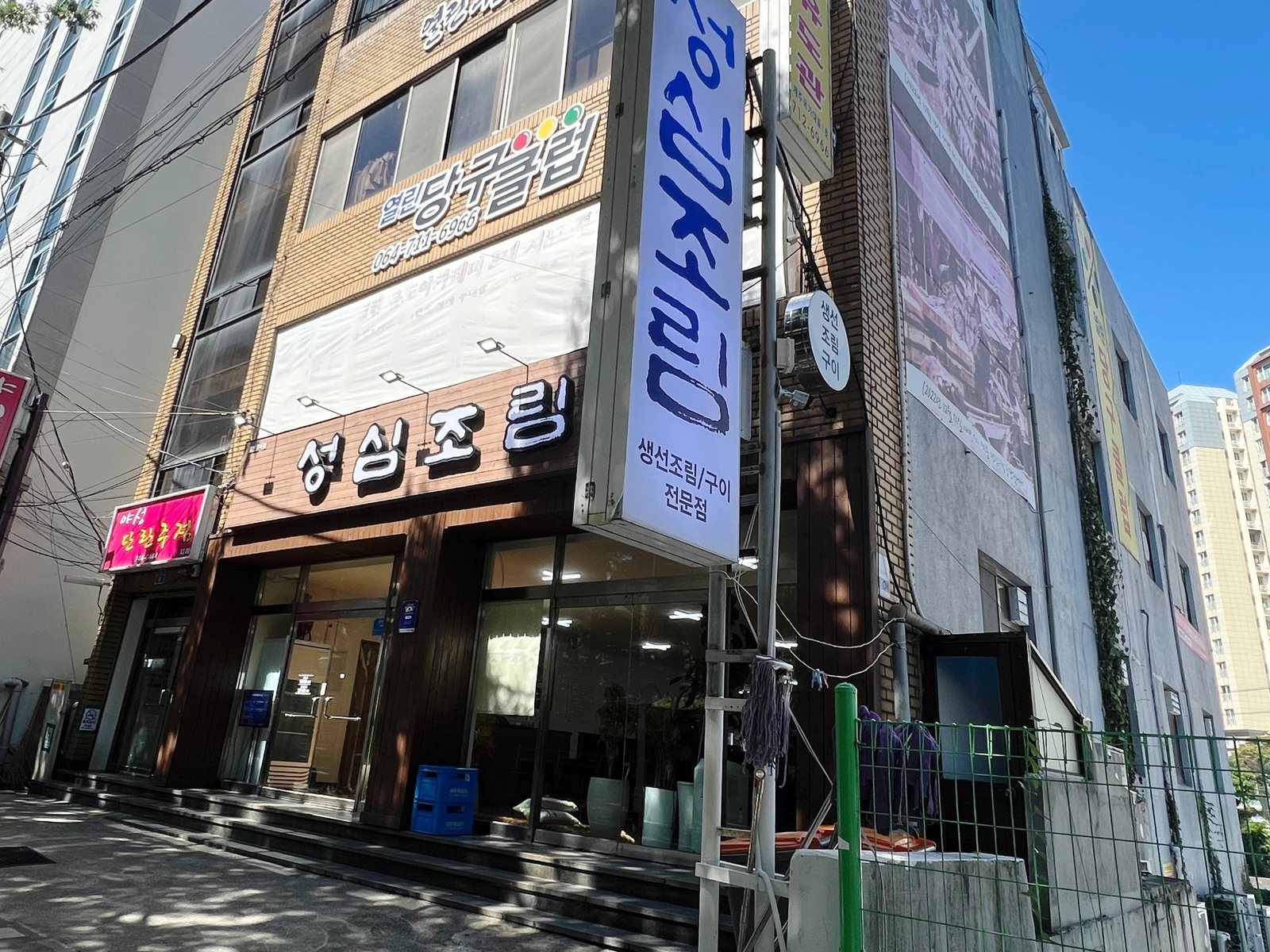 성심조림 제주공항점 외관 — 맑은 날 본 건물 모서리, 1층 원목 외벽의 흰 ‘성심조림’ 간판과 흰 바탕에 파란 붓글씨 세로 기둥 간판 ‘성심조림 생선조림/구이 전문점’.