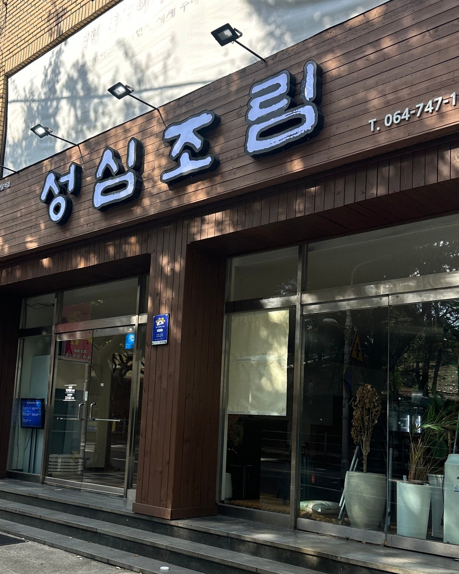 성심조림 제주공항점 외관 — 아침 햇살 아래 짙은 원목 판재 외벽에 흰 붓글씨 입체 간판 ‘성심조림’과 투광등, 그 아래 통유리 출입문과 진열창, 화분.